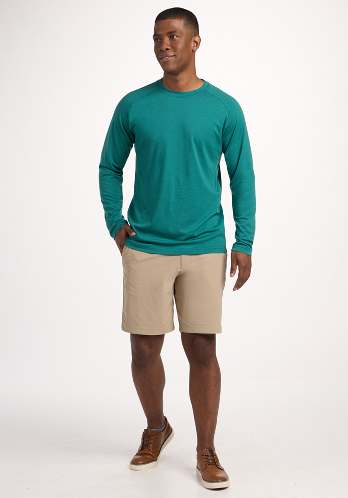 A man stands against a plain white background, wearing the Woolx Essential Tee - Woodland Mini Stripe, beige shorts, and brown shoes. With one hand in his pocket, he smiles slightly and looks off to the side.