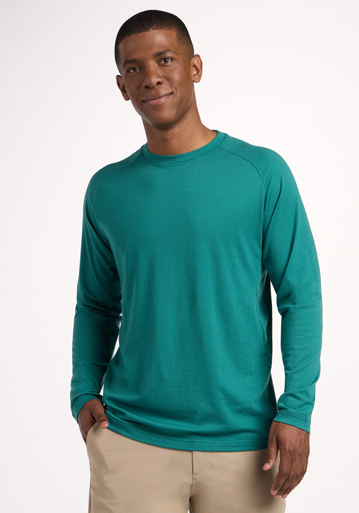 A man smiles against a plain background, wearing the Woolx Essential Tee - Woodland Mini Stripe in teal, paired with beige pants and one hand in his pocket. | Trell is 6’2”, wearing a size M