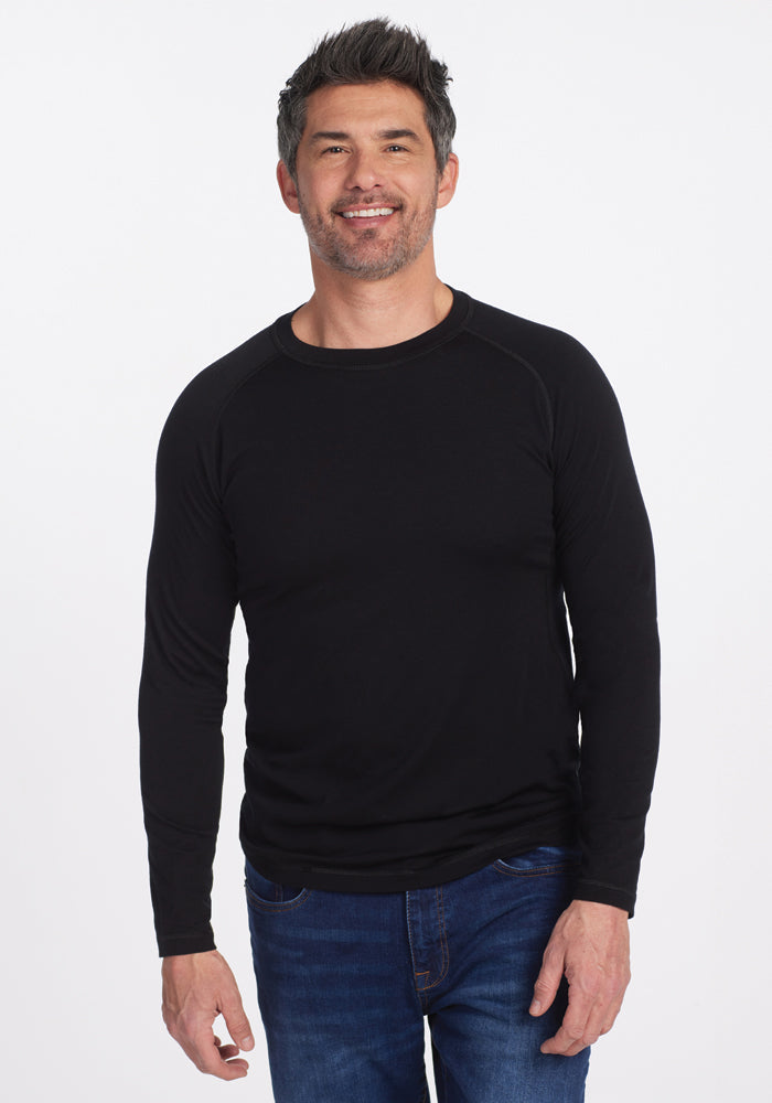 A man with short, dark hair and a beard smiles while wearing a black long-sleeve Essential Tee from Woolx and blue jeans against a plain white background. | Matthew is 6’, wearing a size L