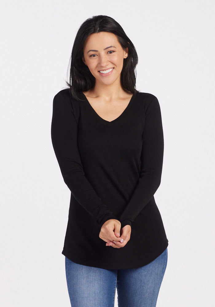 A person with long black hair smiles while wearing the Woolx Eva Tunic in black, a luxuriously soft and warm midweight V-neck long-sleeve shirt, paired with blue jeans against a plain white background, with their hands clasped in front.