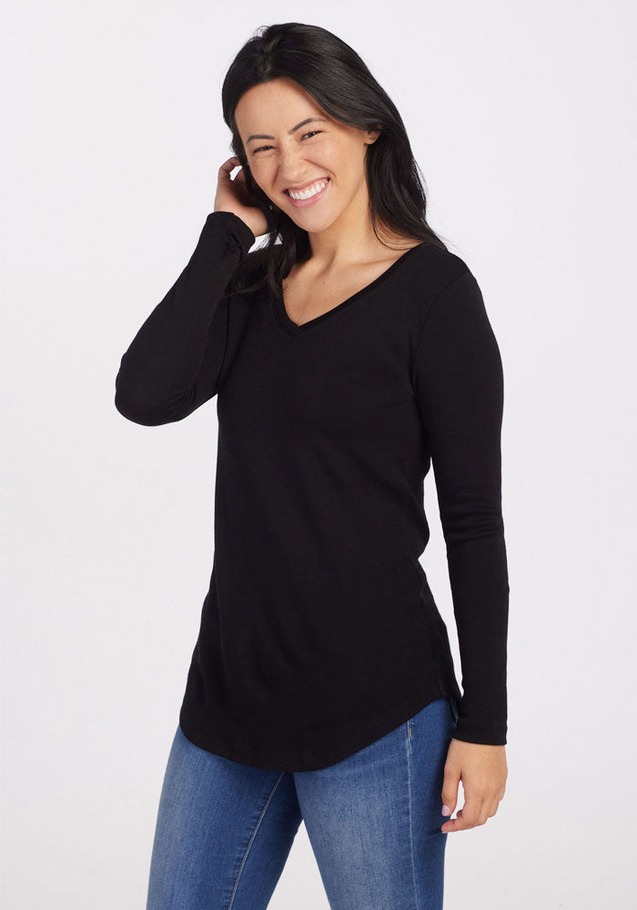 A person with long black hair is smiling and holding their hand to their head. They are wearing a luxuriantly soft, midweight warmth Eva Tunic - Black made of Merino wool from Woolx, paired with blue jeans against a plain white background.