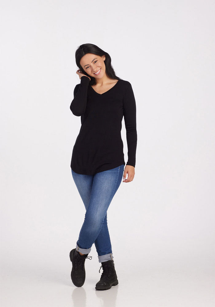 A person with long dark hair is smiling while wearing the luxuriously soft Woolx Eva Tunic in black, crafted from Merino wool. It is paired with blue jeans and black boots. They stand against a plain white background, one hand gently touching their face and the other resting by their side.