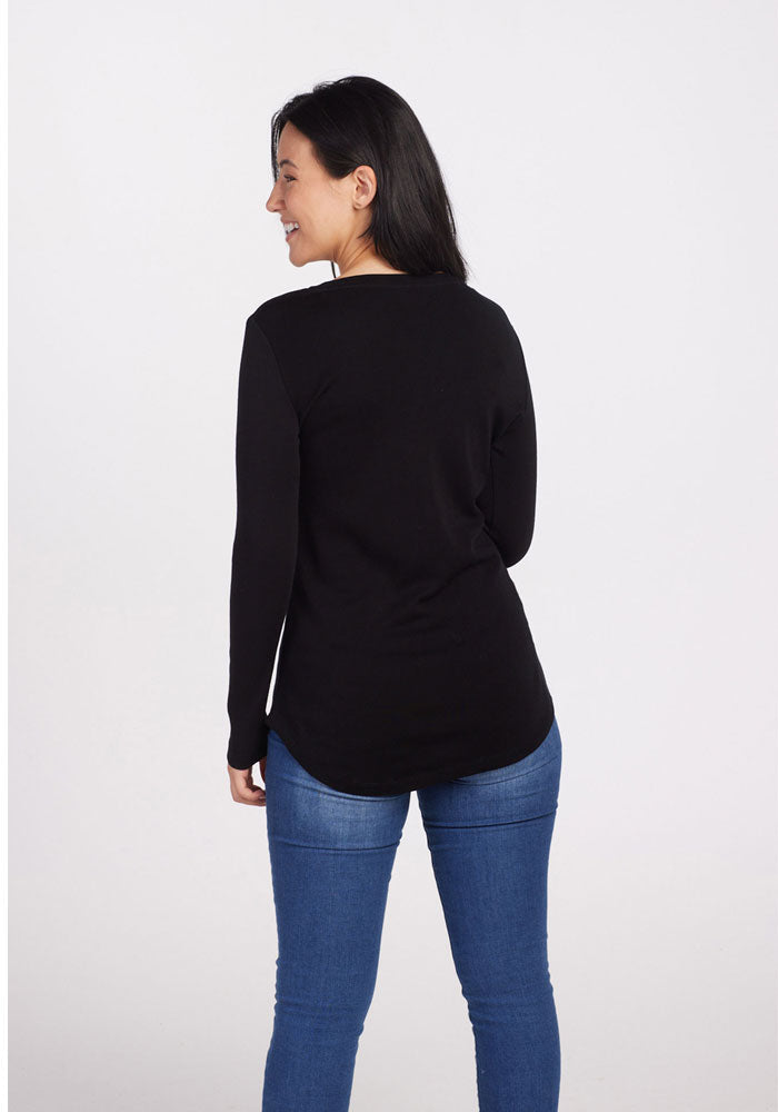 A person with long dark hair, wearing a black Eva Tunic by Woolx and blue jeans, stands smiling and facing slightly to the side against a plain white background. The luxuriously soft Merino wool fabric offers midweight warmth, adding comfort to their stylish appearance.