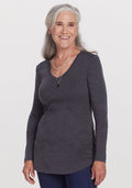 Smiling older woman with long gray hair, wearing the Woolx Eva Tunic in Cinder Melange, blue jeans, and layered necklaces, stands against a plain white background. | Anne is 5’8”, wearing a size S