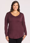 A woman with long brown hair, wearing the Woolx Eva Tunic in Chocolate Fig and black pants, stands smiling against a plain light background.
