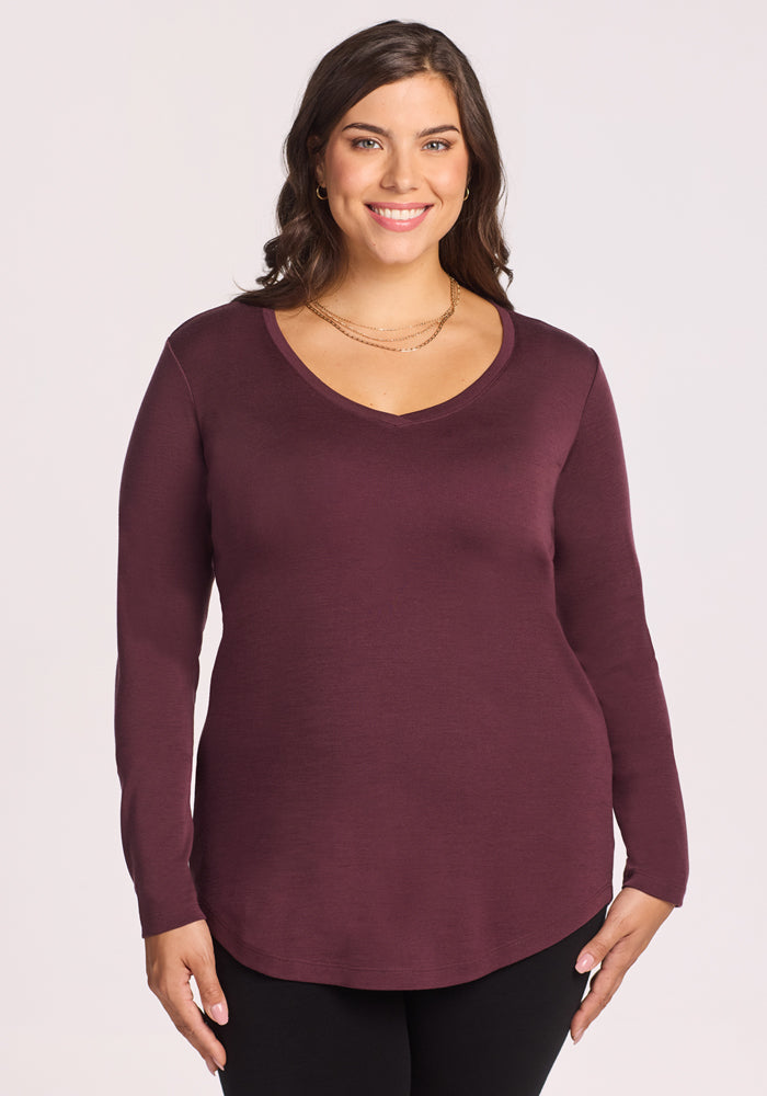 A woman with long brown hair, wearing the Woolx Eva Tunic in Chocolate Fig and black pants, stands smiling against a plain light background.