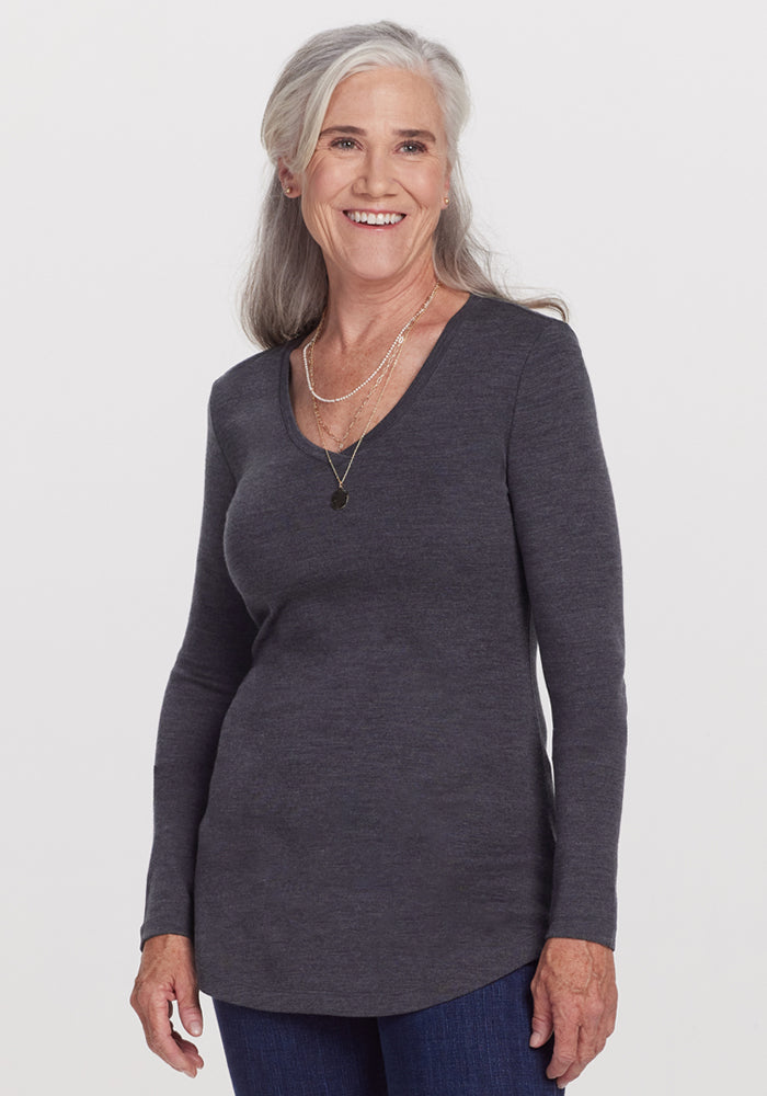 Smiling older woman with long gray hair, wearing the Woolx Eva Tunic in Cinder Melange, blue jeans, and layered necklaces, stands against a plain white background. | Anne is 5’8”, wearing a size S
