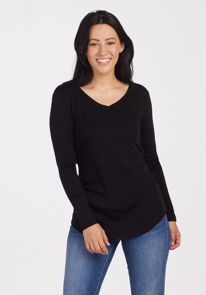 A person smiling, wrapped in the midweight warmth of a black Woolx Eva Tunic and blue jeans. They have luxuriously soft, long dark hair and are standing against a plain white background.