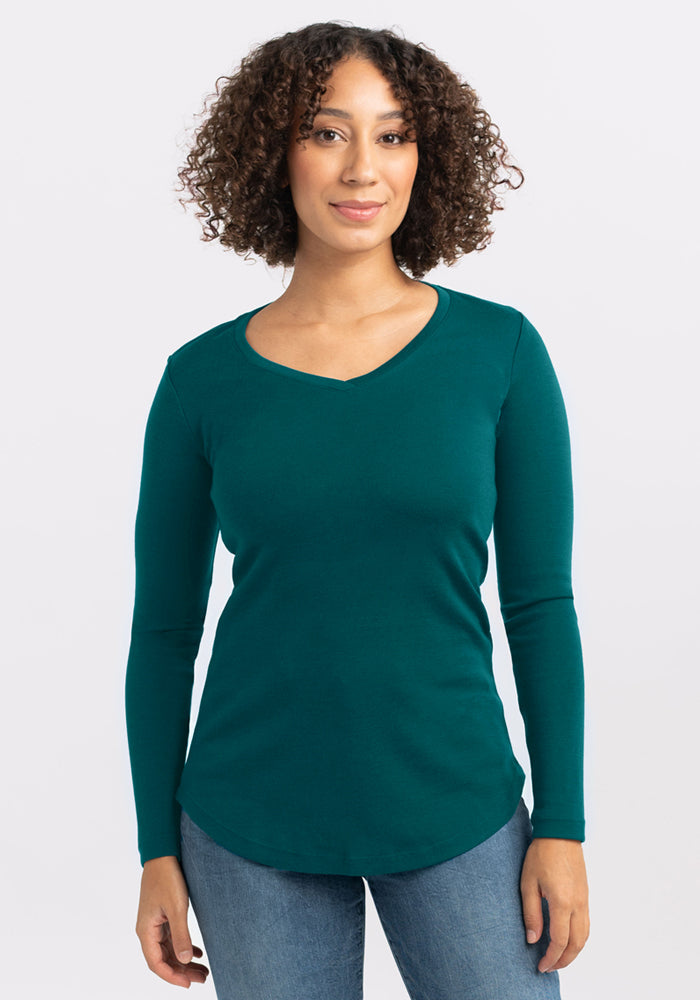 A woman with curly hair wears the Woolx Eva Tunic in Riverbank Green and blue jeans, standing facing the camera against a plain light background.