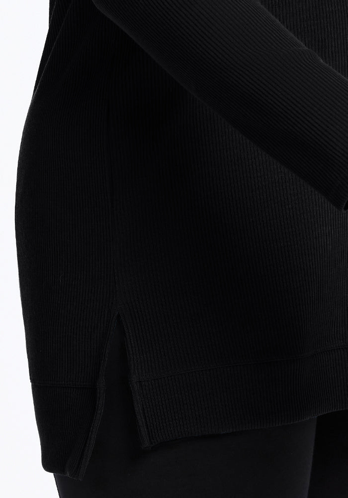 The close-up image features an individual wearing the Evie Tunic in black by Woolx, a temperature-regulating ribbed sweater with a side slit. Made from Merino wool, this comfortable tunic is styled with black pants, and its textured fabric accentuates the sweater's chic design.