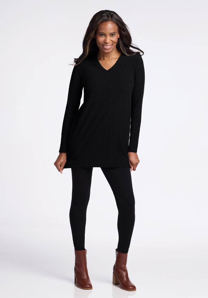 A person with long hair smiles while wearing the Evie Tunic in black from Woolx, crafted from temperature-regulating Merino wool, paired with black leggings and brown ankle boots. They stand against a plain white background.
