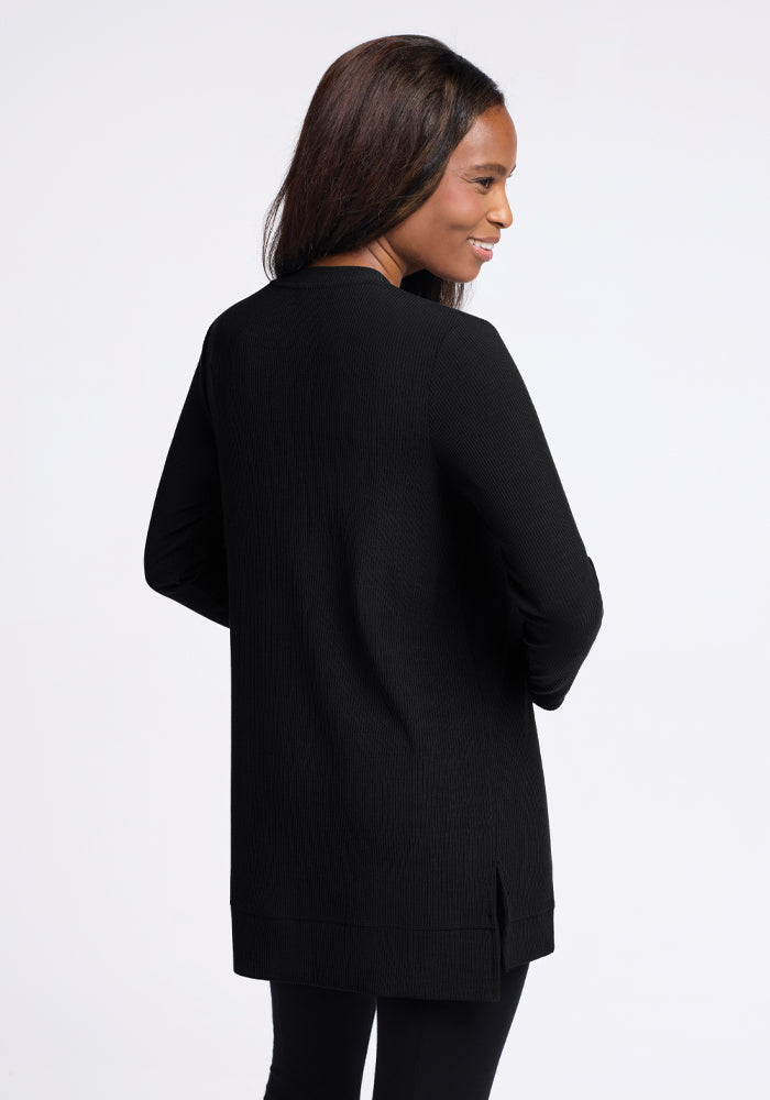 A woman with long hair is wearing the cozy, flattering Evie Tunic in black from Woolx. She is smiling and slightly facing to the right, standing against a plain, light background.
