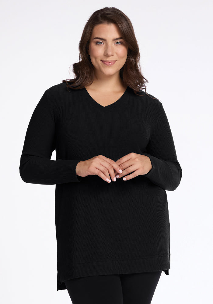 A person with long brown hair smiles at the camera, wearing a cozy black Evie Tunic by Woolx, made from temperature-regulating Merino wool. They have a relaxed pose with hands gently clasped in front of them against a plain white background.