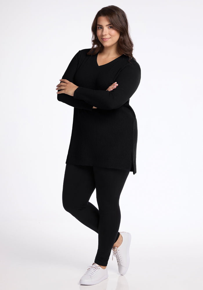 A person stands confidently with arms crossed, wearing the Evie Tunic - Black by Woolx along with matching leggings. They are smiling in white sneakers. This cozy tunic, set against a plain white background, is perfect for any season due to its temperature-regulating qualities.