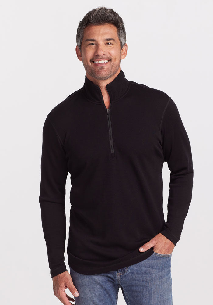 A person with short, gray hair is smiling and standing against a plain white background. They are wearing an ultra-soft Explorer 1/4 Zip pullover in black from Woolx, made of midweight fabric, paired with blue jeans. One hand is in their pocket while the other rests at their side. | Matthew is 6’, wearing a size L
