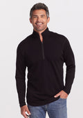 A person with short, gray hair is smiling and standing against a plain white background. They are wearing an ultra-soft Explorer 1/4 Zip pullover in black from Woolx, made of midweight fabric, paired with blue jeans. One hand is in their pocket while the other rests at their side. | Matthew is 6’, wearing a size L