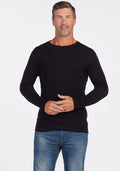 A man with short, slightly wavy hair is standing against a plain white background. He is wearing a black long-sleeve Explorer Base Layer from Woolx and blue jeans. He has a neutral expression and is facing the camera, with his hands loosely clasped in front of him. | Lee is 6’, wearing a size M