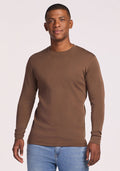 A man stands facing the camera against a plain light background, smiling slightly, wearing the Woolx Explorer Base Layer • Final Sale - Bourbon Oak and light blue jeans.