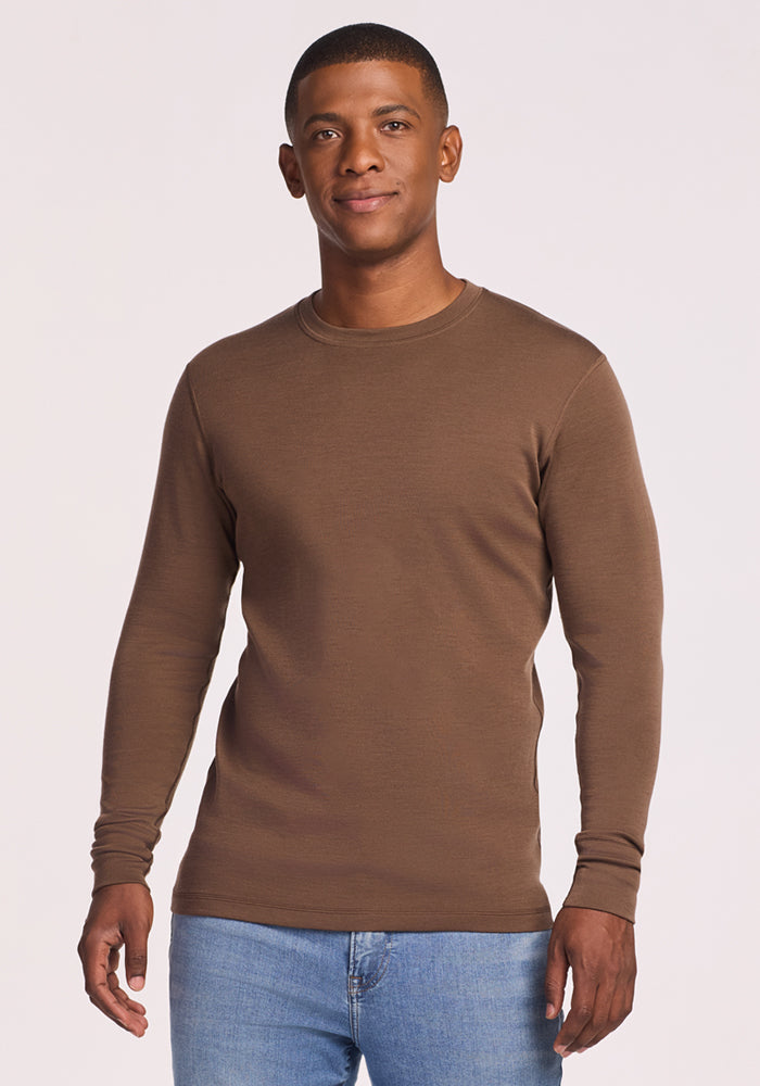 A man stands facing the camera against a plain light background, smiling slightly, wearing the Woolx Explorer Base Layer • Final Sale - Bourbon Oak and light blue jeans.