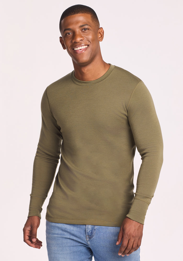 A smiling man wears the Woolx Explorer Base Layer in Winter Moss with light blue jeans, standing against a plain light background.