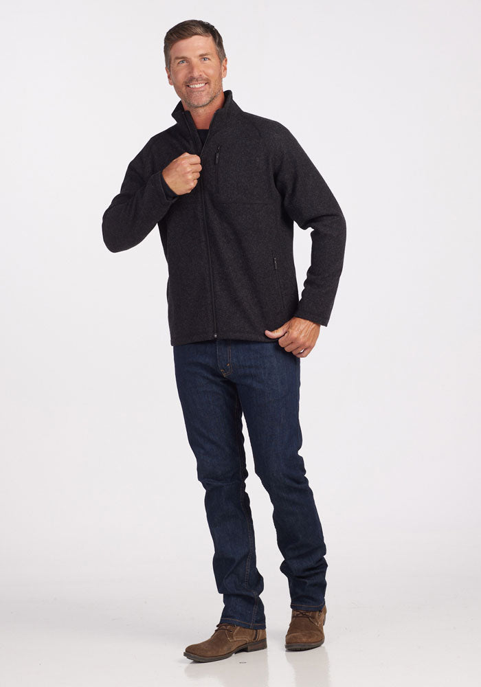 A man wearing the Fairbanks Jacket OLD by Woolx, a minimalistic dark zip-up crafted from merino wool for optimal insulation, stands against a plain white background. He pairs it with blue jeans and brown shoes and is smiling as he holds the open collar of his jacket with one hand.
