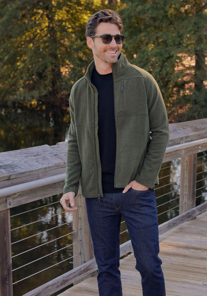 A man in the Woolx Fairbanks Jacket - Dark Forest, sunglasses, and dark jeans stands smiling on a wooden bridge, surrounded by trees and water on a sunny day.