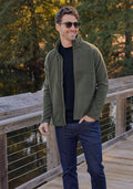 A man in the Woolx Fairbanks Jacket - Dark Forest, sunglasses, and dark jeans stands smiling on a wooden bridge, surrounded by trees and water on a sunny day.