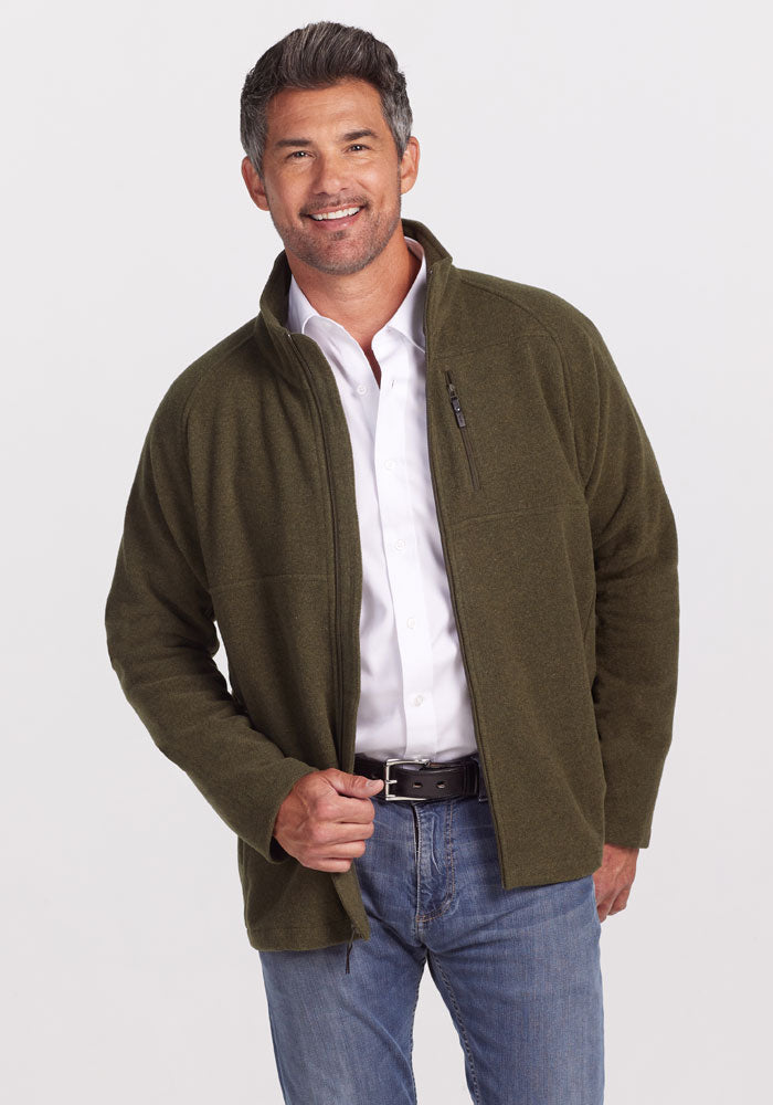 A man wearing a white shirt and an olive green Fairbanks Jacket by Woolx stands smiling with one hand in his jeans pocket, set against a plain gray background. | Matthew is 6', wearing a size L