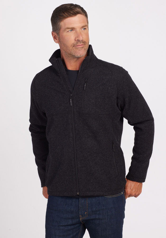 A man with short hair is wearing a Woolx Fairbanks Jacket in Carbon Black, featuring a zip-up design with a minimalistic aesthetic. Paired with jeans, he stands against a plain background, looking to the side with one hand in his pocket, highlighting the refined simplicity of his attire. | Lee is 6’, wearing a size M