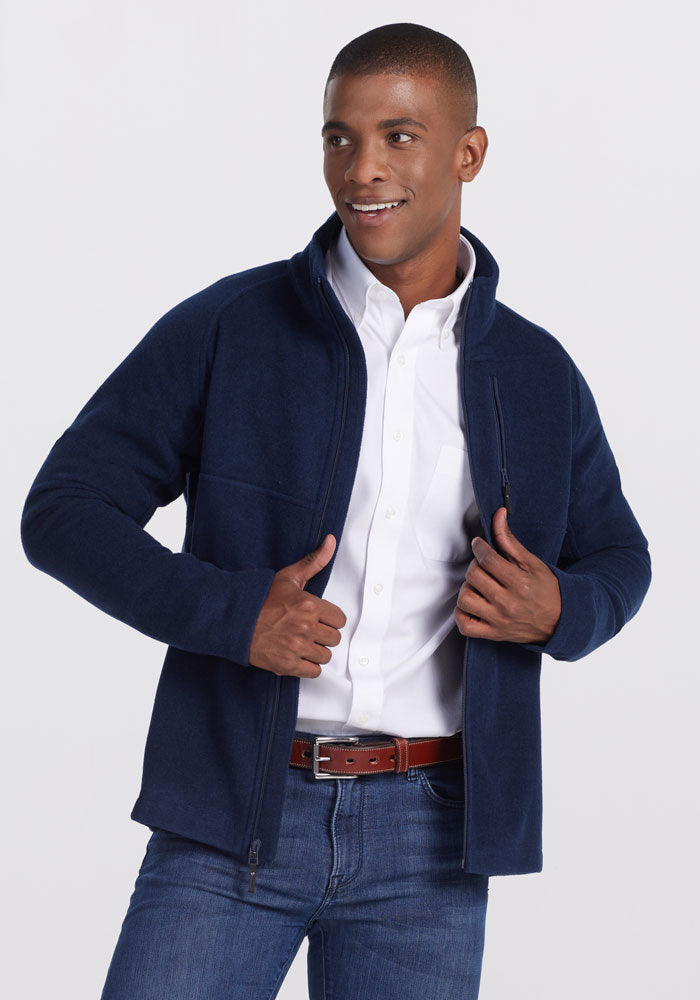 A person wearing a dark navy Fairbanks Jacket from Woolx over a white button-up shirt and blue jeans poses confidently. They are smiling and adjusting their ultra-heavyweight jacket with both hands against a plain, light-colored background. | Trell is 6’2”, wearing a size M