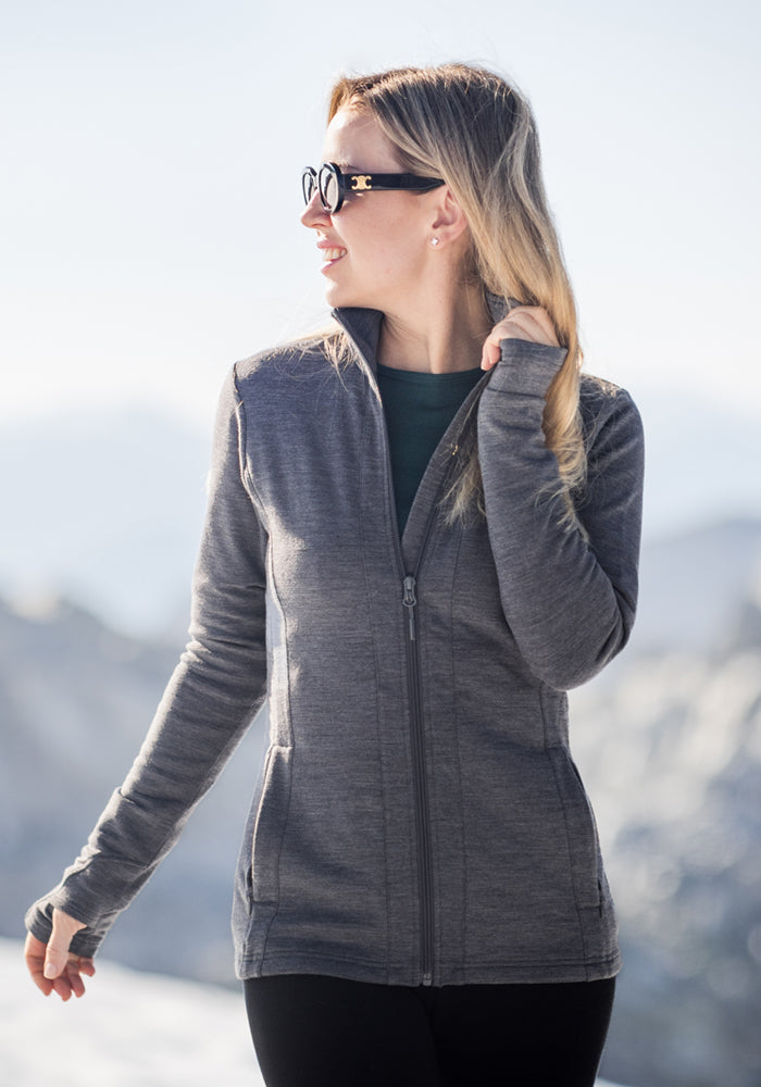 A woman in a Woolx Finley Zip Up Sweatshirt and sunglasses stands outdoors in the mountains, smiling and looking to the side against a bright, softly blurred background.