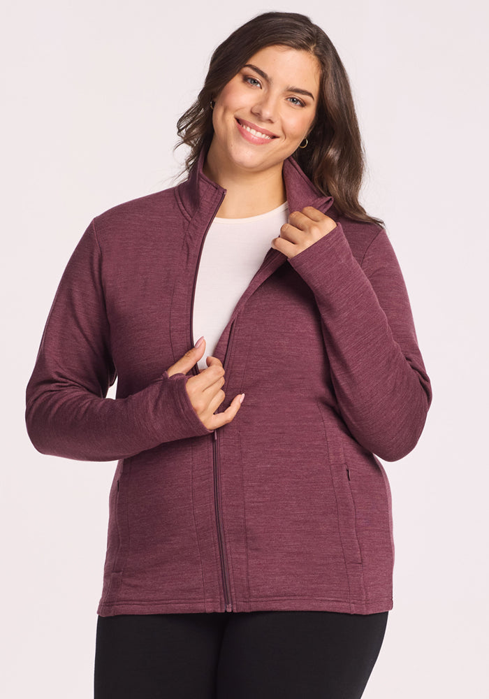 A woman with long brown hair smiles while wearing the Woolx Finley Zip Up Sweatshirt in Grape Harvest Melange over a white top, holding the collar and zipper against a plain light background.