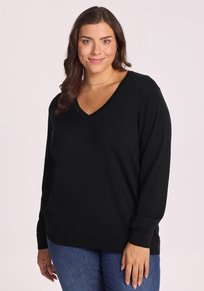 A woman with long brown hair smiles softly at the camera, wearing the travel-friendly Woolx Francesca Sweater in black and blue jeans, standing against a plain light background. | Juliana is 5'10", wearing a size XL