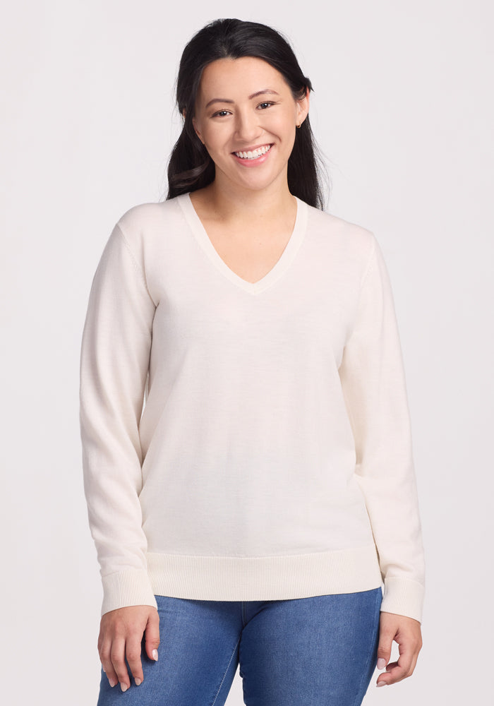 A smiling person with long dark hair is wearing the Woolx Francesca Sweater in Dewdrop Pearl and blue jeans against a plain background. This cozy, travel-friendly sweater blends style with breathable comfort for any adventure. | Sarah is 5’8”, wearing a size S