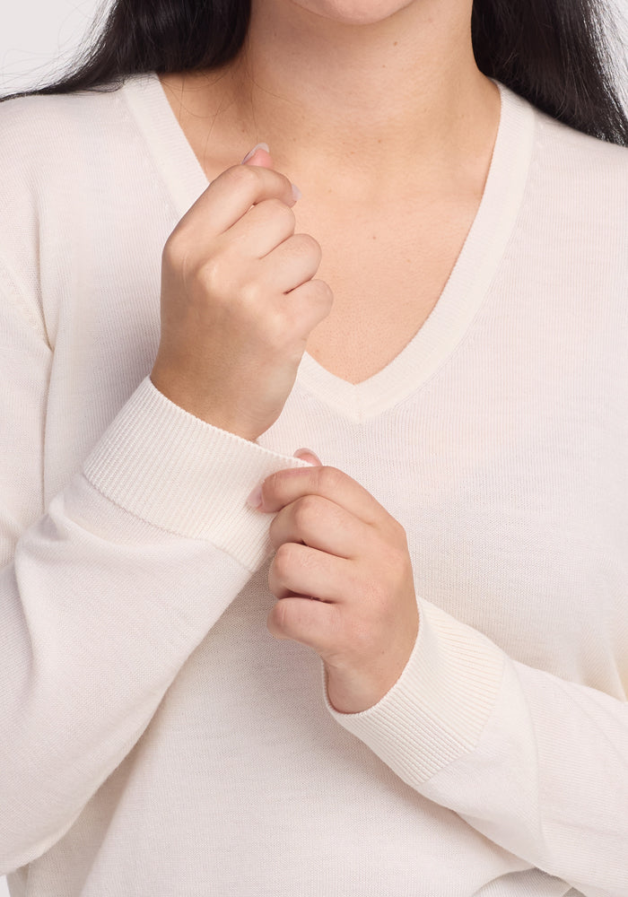 A person wearing the Francesca Sweater - Dewdrop Pearl by Woolx, a cream long-sleeve V-neck made from breathable Merino wool, casually tugs at the sleeve while relaxing the other arm, showcasing its travel-friendly style.