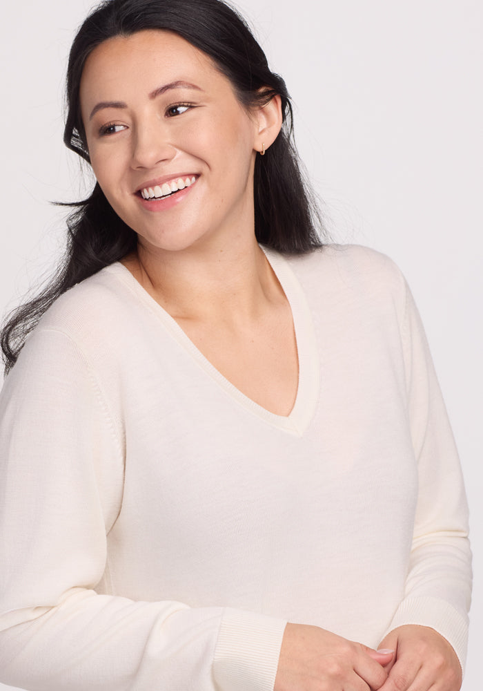 A person with long dark hair smiles, looking to the side while wearing the Francesca Sweater - Dewdrop Pearl by Woolx. Made from breathable Merino wool, this light-colored V-neck sweater stands out against the plain background, enhancing its travel-friendly appeal.