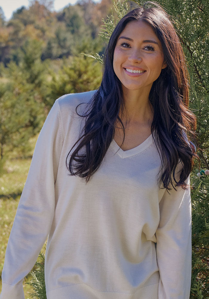 A woman with long dark hair smiles outdoors among green trees on a sunny day, wearing the Woolx Francesca Sweater in Dewdrop Pearl.