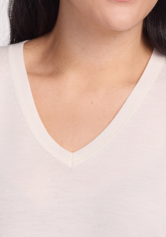 A close-up features someone wearing Woolx's Francesca Sweater in Dewdrop Pearl. The white V-neck, made from soft Merino wool, highlights the neckline and part of the chest without showing facial features. This lightweight, moisture-wicking fabric makes it perfect for travel.