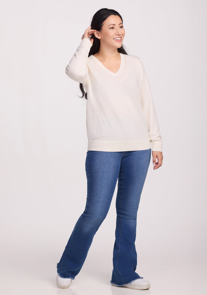 A person with long dark hair smiles, gazing to the side. They wear a Woolx Francesca Sweater in Dewdrop Pearl, perfect for travel with its breathable Merino wool. Complimented by blue jeans and white sneakers, they stand against a plain white background.