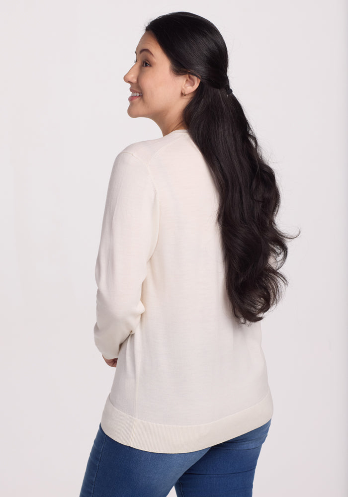 A person with long, wavy black hair smiles over their shoulder, wearing the travel-friendly Francesca Sweater in Dewdrop Pearl by Woolx and blue jeans against a plain white background.