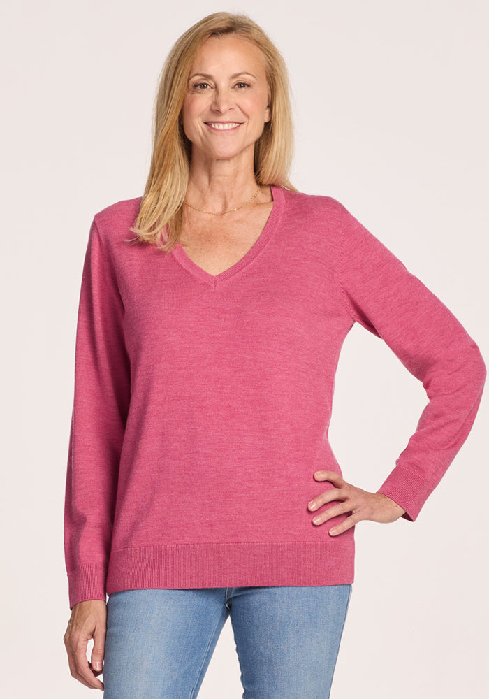 A smiling woman with straight blonde hair wears the Woolx Francesca Sweater in Wild Salmon and light blue jeans, standing against a plain light background with one hand on her hip. | Andrea is 5’10”, wearing a size S