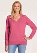A smiling woman with straight blonde hair wears the Woolx Francesca Sweater in Wild Salmon and light blue jeans, standing against a plain light background with one hand on her hip. | Andrea is 5’10”, wearing a size S