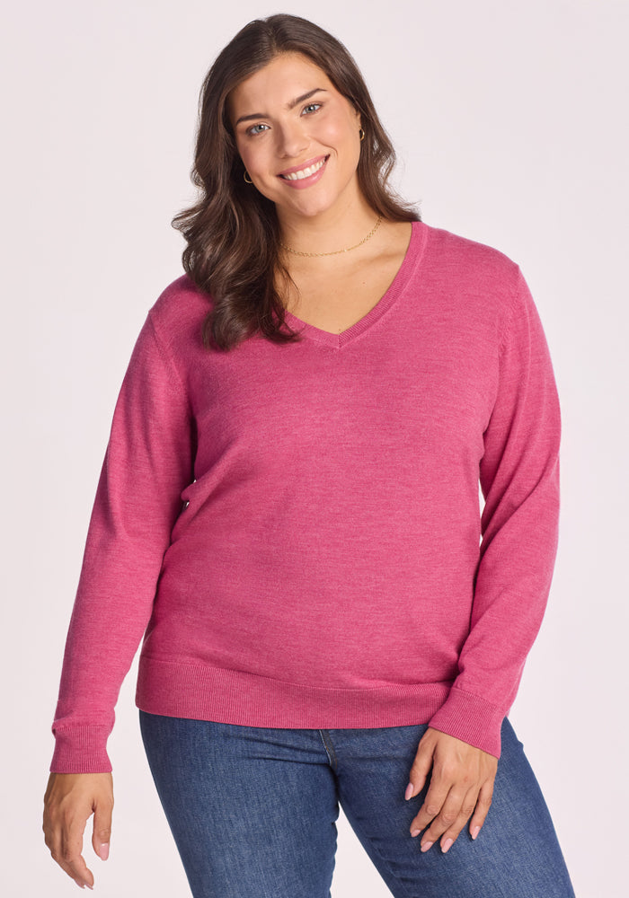 A woman with long brown hair, wearing the Woolx Francesca Sweater in Wild Salmon and blue jeans, smiles while standing against a plain light background. | Juliana is 5'10", wearing a size XL