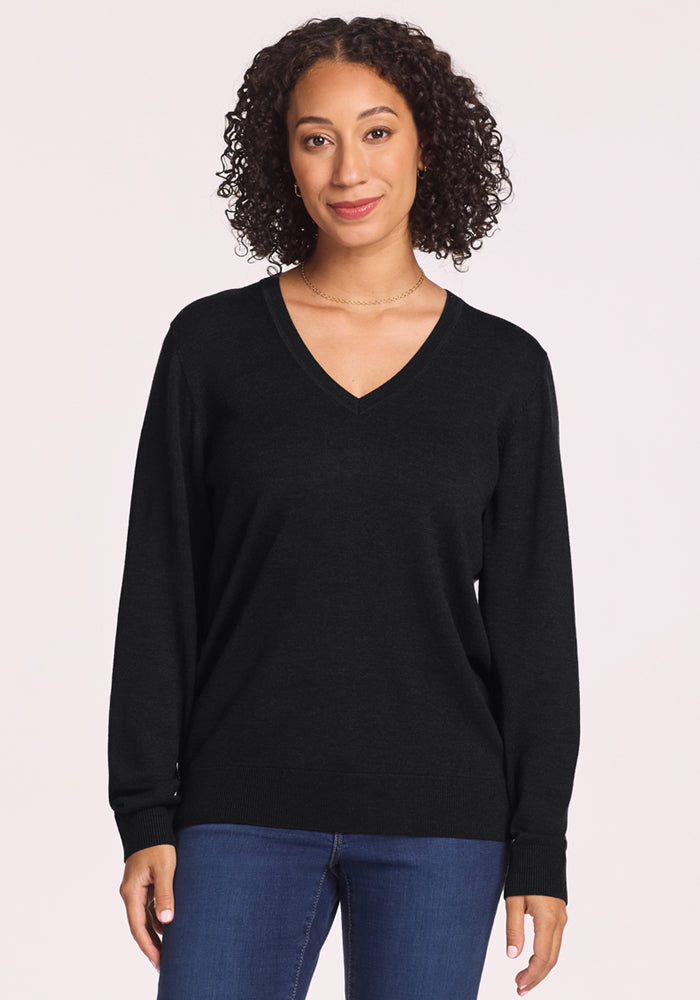 A woman with curly hair wears the Woolx Francesca Sweater in black and blue jeans. Against a plain light background, she smiles slightly at the camera, effortlessly showcasing this travel-friendly sweater. | Tori is 5'7", wearing a size S