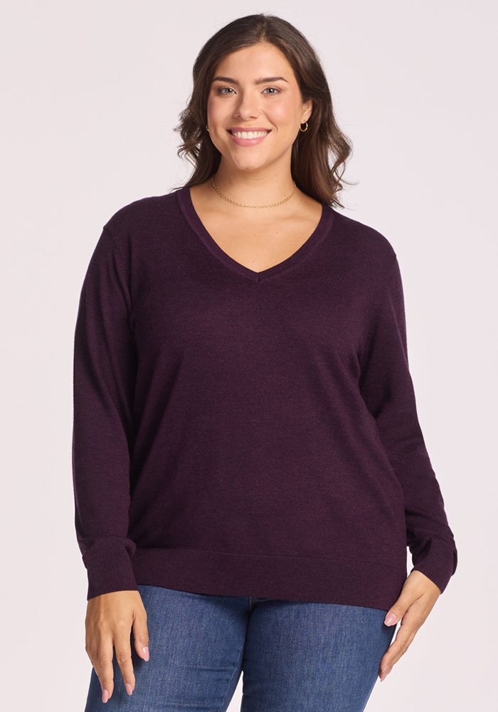 A woman with long brown hair smiles, wearing the Woolx Francesca Sweater in Deep Plum and blue jeans, standing against a plain light background.