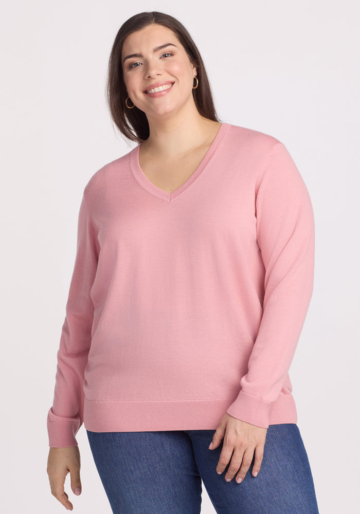 A woman with long brown hair smiles while wearing the Woolx Francesca Sweater in Blush (Final Sale) and blue jeans, standing against a plain light background.