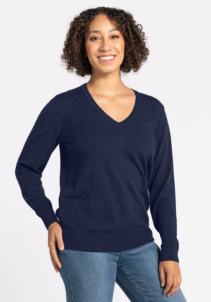 A smiling person with curly hair wears the Woolx Francesca Sweater • Final Sale - Storm Cloud and blue jeans, standing against a plain white background.