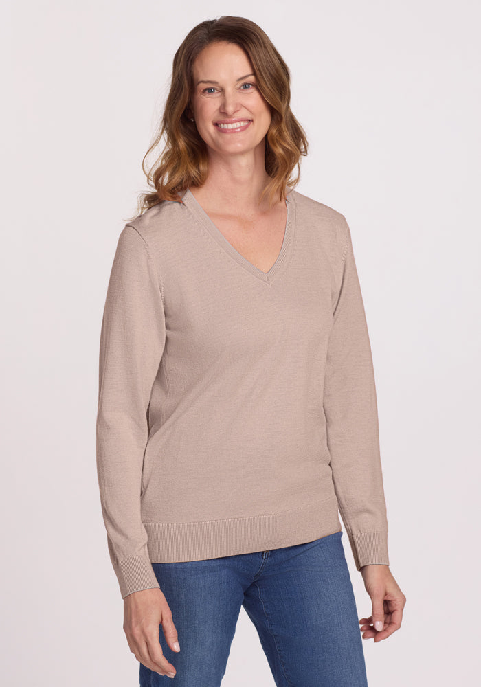 A smiling woman with wavy brown hair wears the Woolx Francesca Sweater in Sandstone Haze and blue jeans, standing against a plain light background.