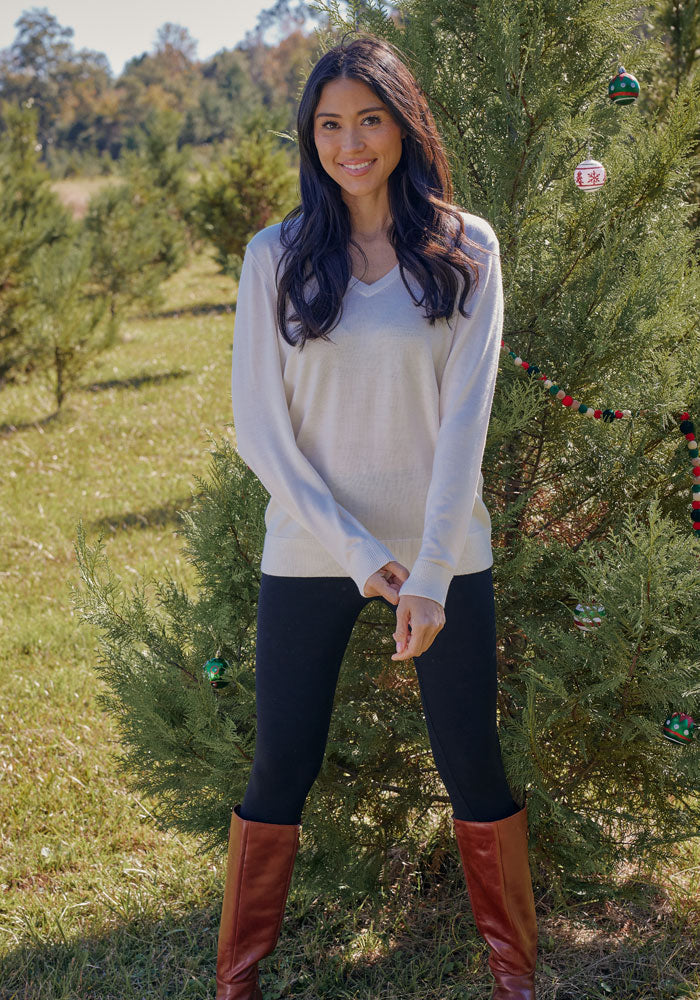 A woman with long dark hair smiles outdoors beside a decorated evergreen, wearing the Woolx Francesca Sweater in Dewdrop Pearl, paired with black leggings and brown boots on a sunny day.