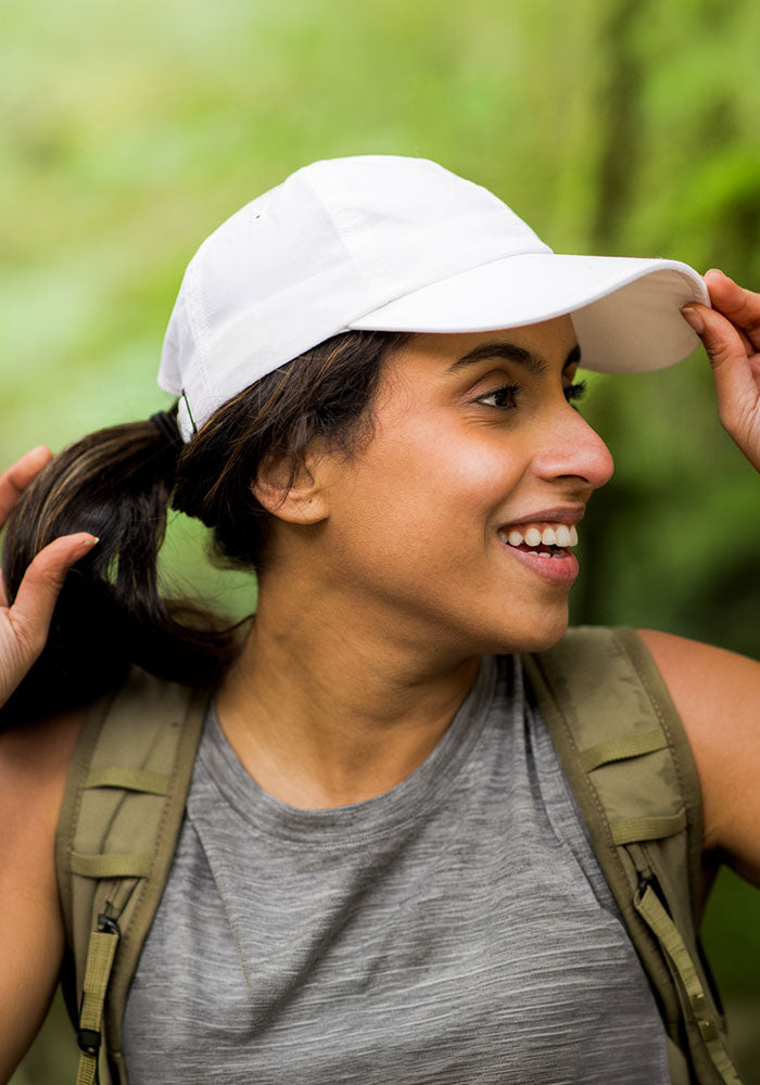 A woman smiles outdoors, wearing the Woolx Frankie Baseball Hat in Polar White, a gray tank top, and a khaki backpack. She holds her ponytail against a lush green backdrop, suggesting she’s hiking or exploring nature.